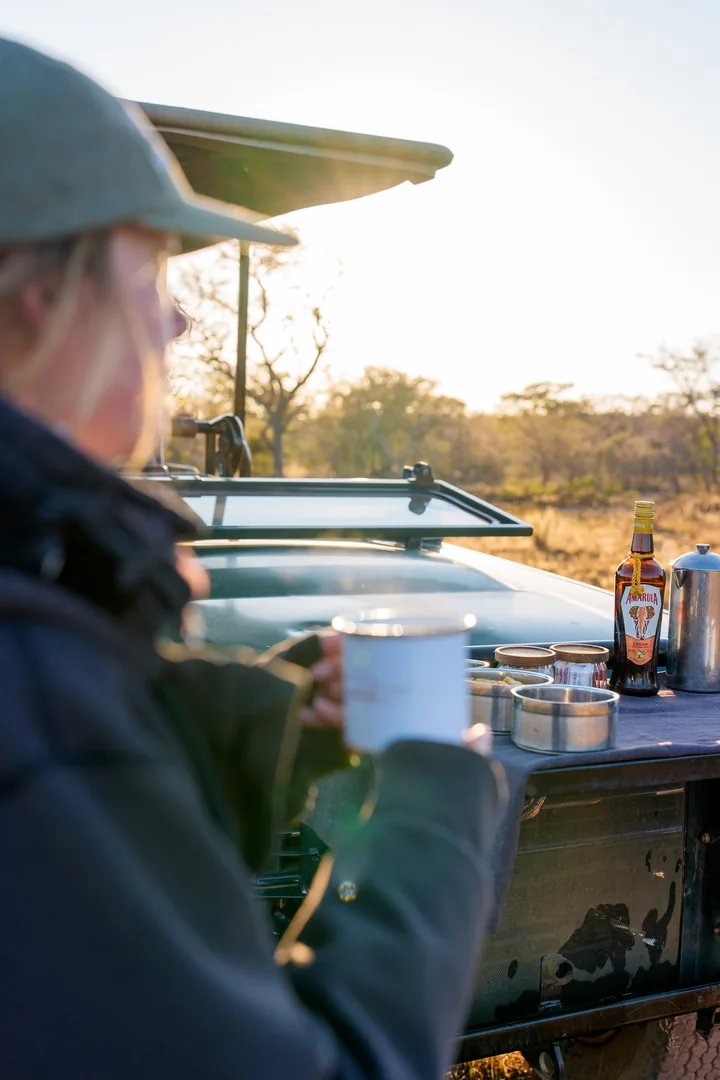A safari guest holding a mug beside a coffee setup on a game drive vehicle.