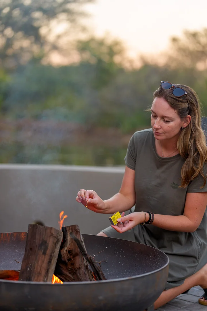 A woman lighting a fire in an outdoor fire bowl at sunset.